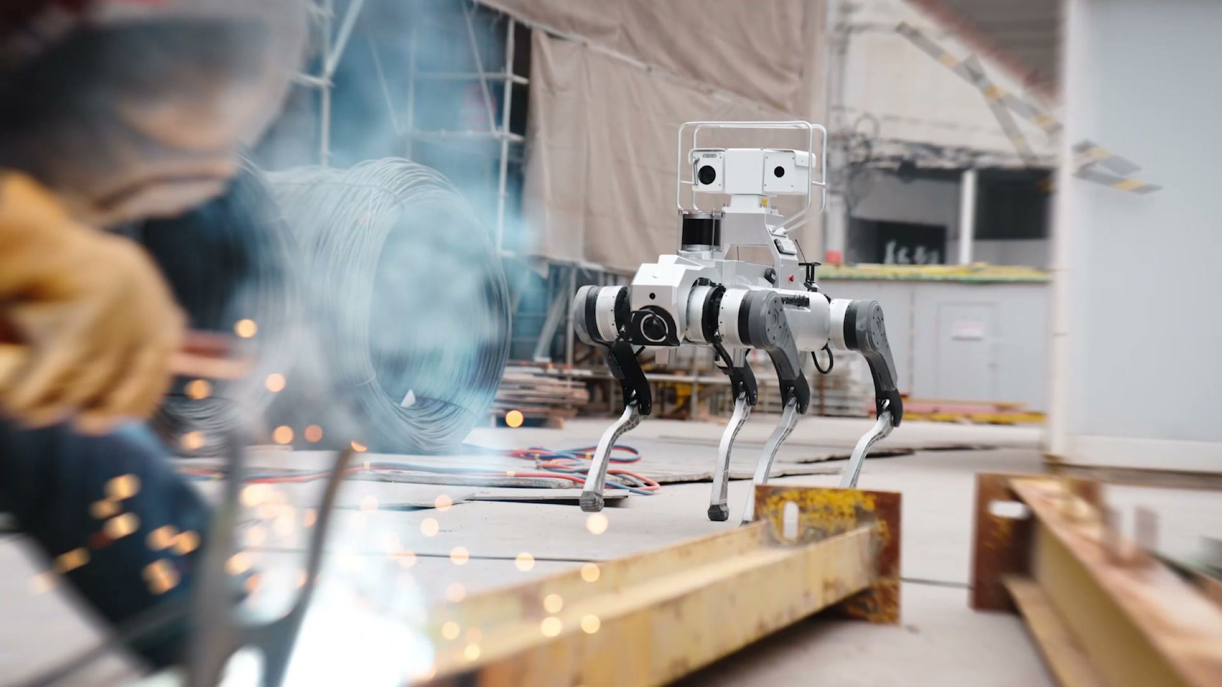 A quadruped robot carrying a camera and sensor mast patrols a worksite while a welder works in the foreground.