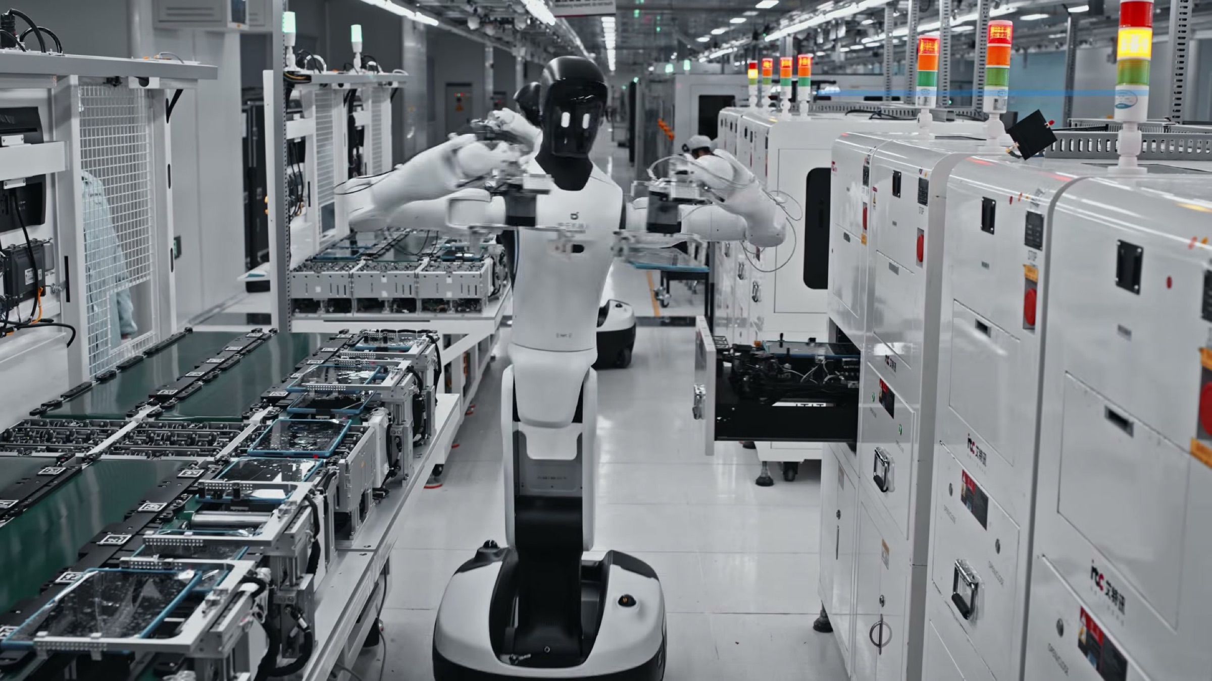 A wheeled dual-arm humanoid loads devices into a test station on a live electronics production line.