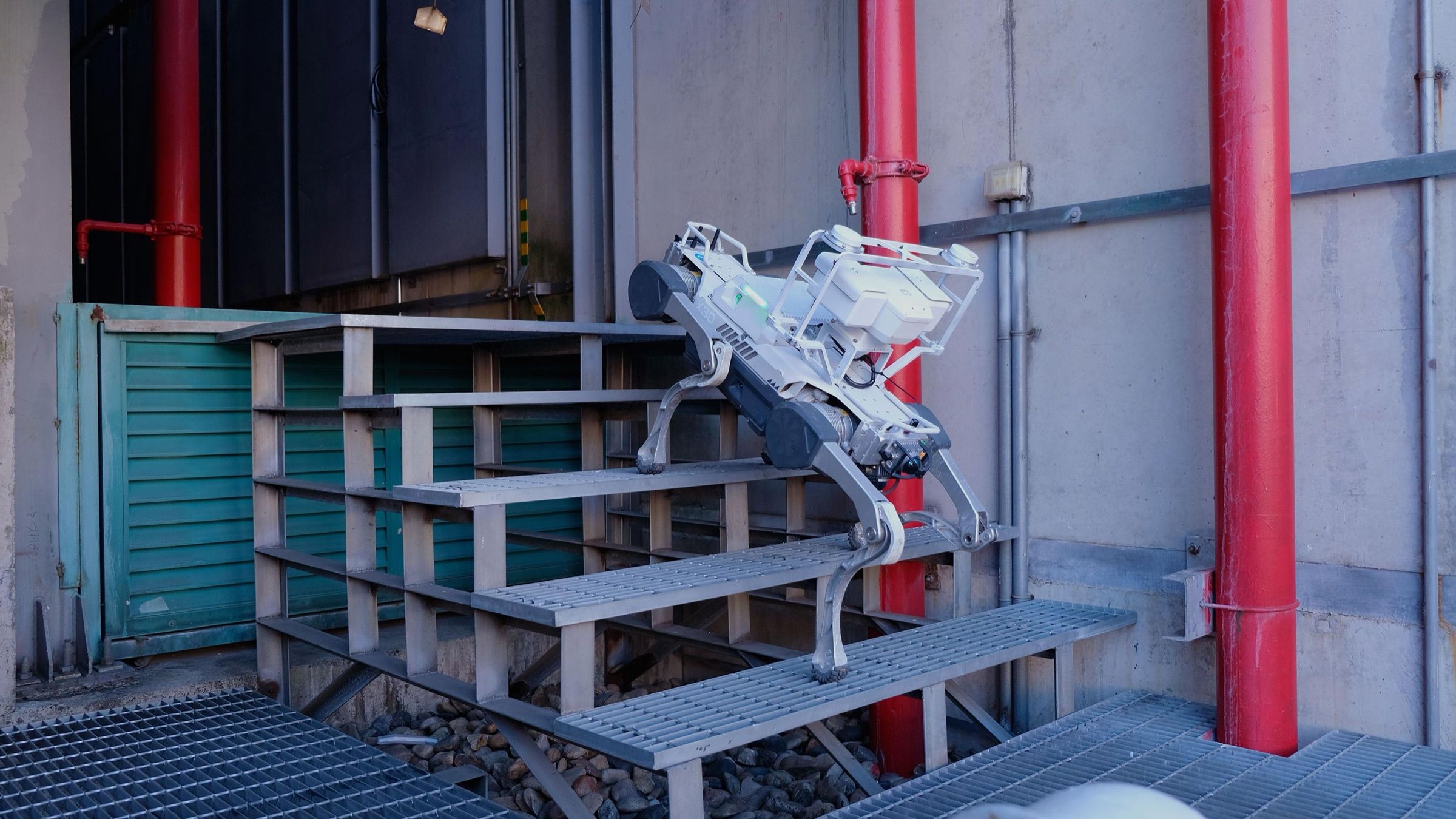 A quadruped inspection robot carrying sensor payloads climbs steel grating stairs beside red fire-water pipes at an industrial power facility.
