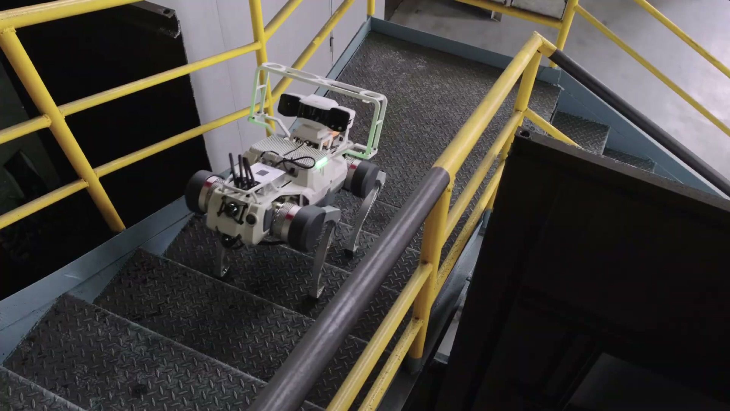 A quadruped robot with a camera and sensor frame descends a yellow-railed steel staircase inside a heavy industrial plant (frame at 0:28).