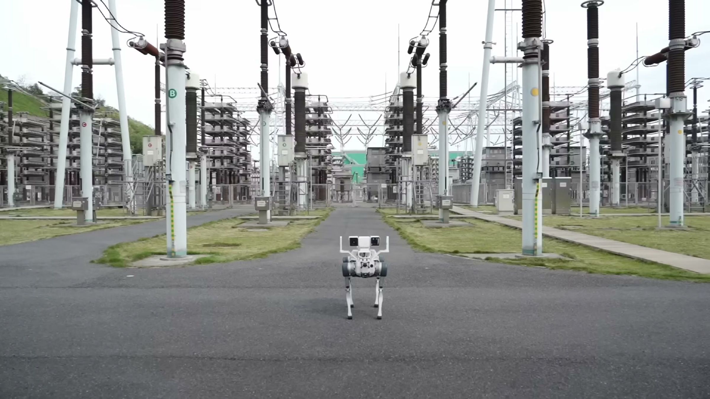 A quadruped robot with a camera mast walks an access road between high-voltage switchgear at an electrical substation (frame at 0:21.5).