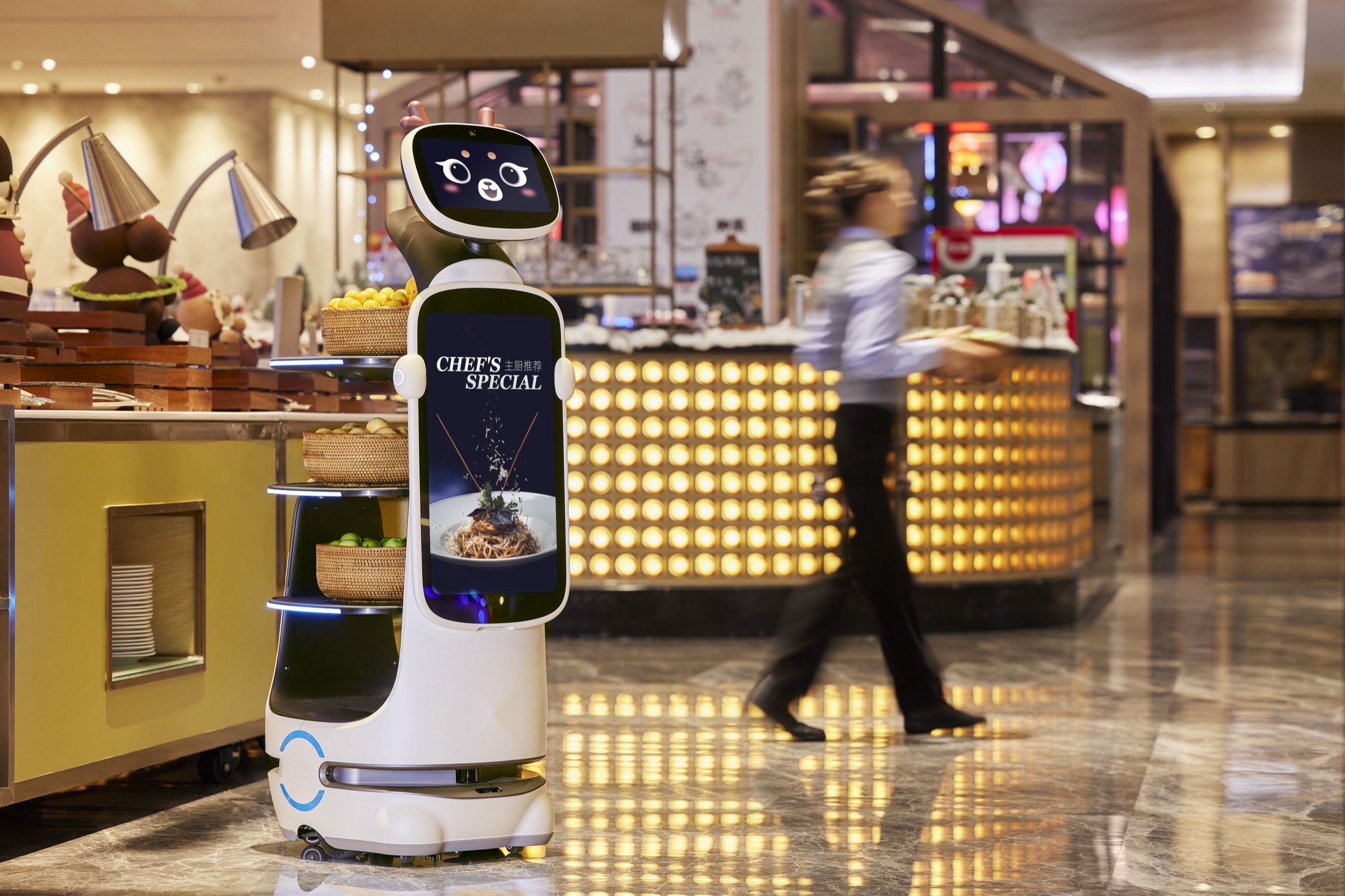 Service robot carrying a dish past a buffet counter as a staff member walks by in a hotel restaurant