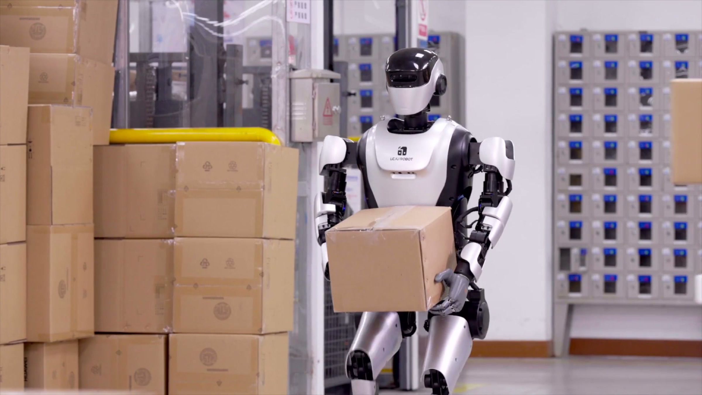 A humanoid robot carries a carton past stacked boxes in a factory storeroom. Frame at 47.5s.