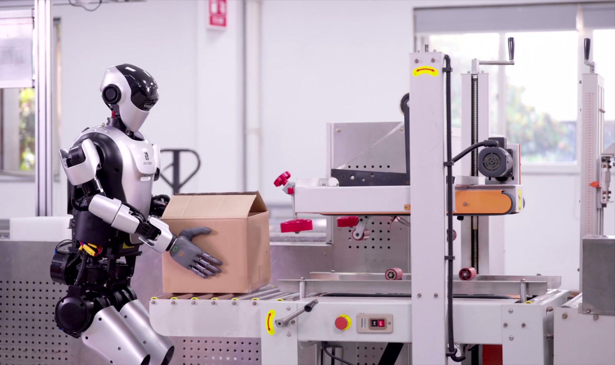 A humanoid robot places a cardboard carton onto a carton-taping machine on a packing line. Frame at 34.5s, small edge crop.