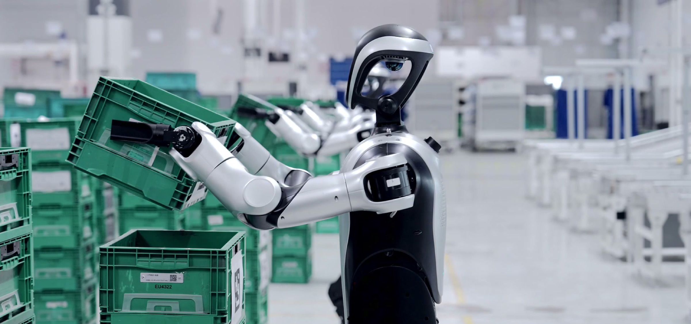 A wheeled humanoid robot lifts green plastic totes off a stack in a parts logistics area. Frame at 4.0s, cropped to remove a burned-in caption.