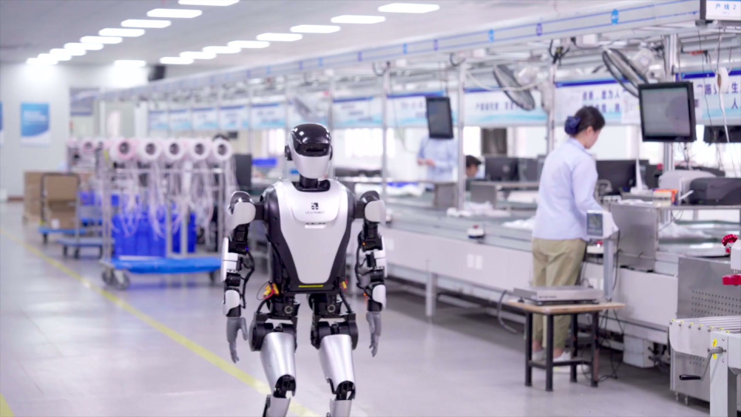 A humanoid robot walks along a production aisle while staff work at assembly stations. Frame at 0.8s.