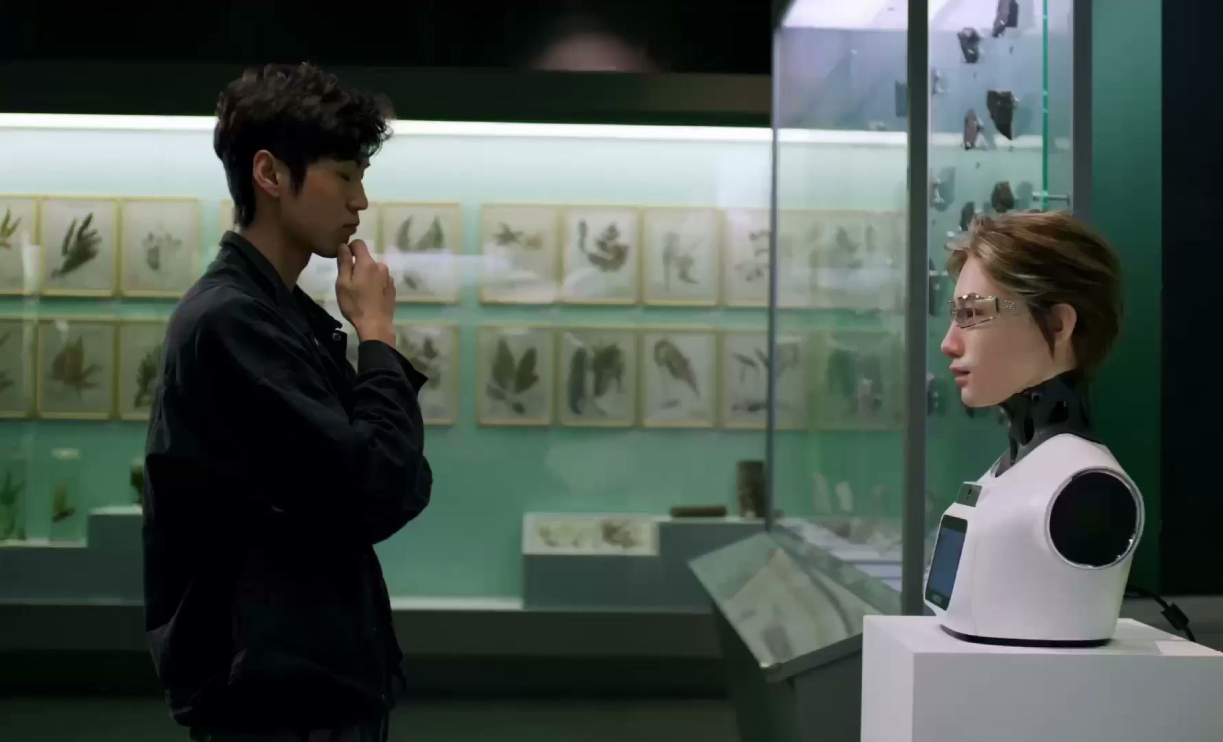 A lifelike desktop robot answers a visitor’s questions beside a specimen display in an exhibition hall. Frame at 18.5s, cropped to remove a text panel.