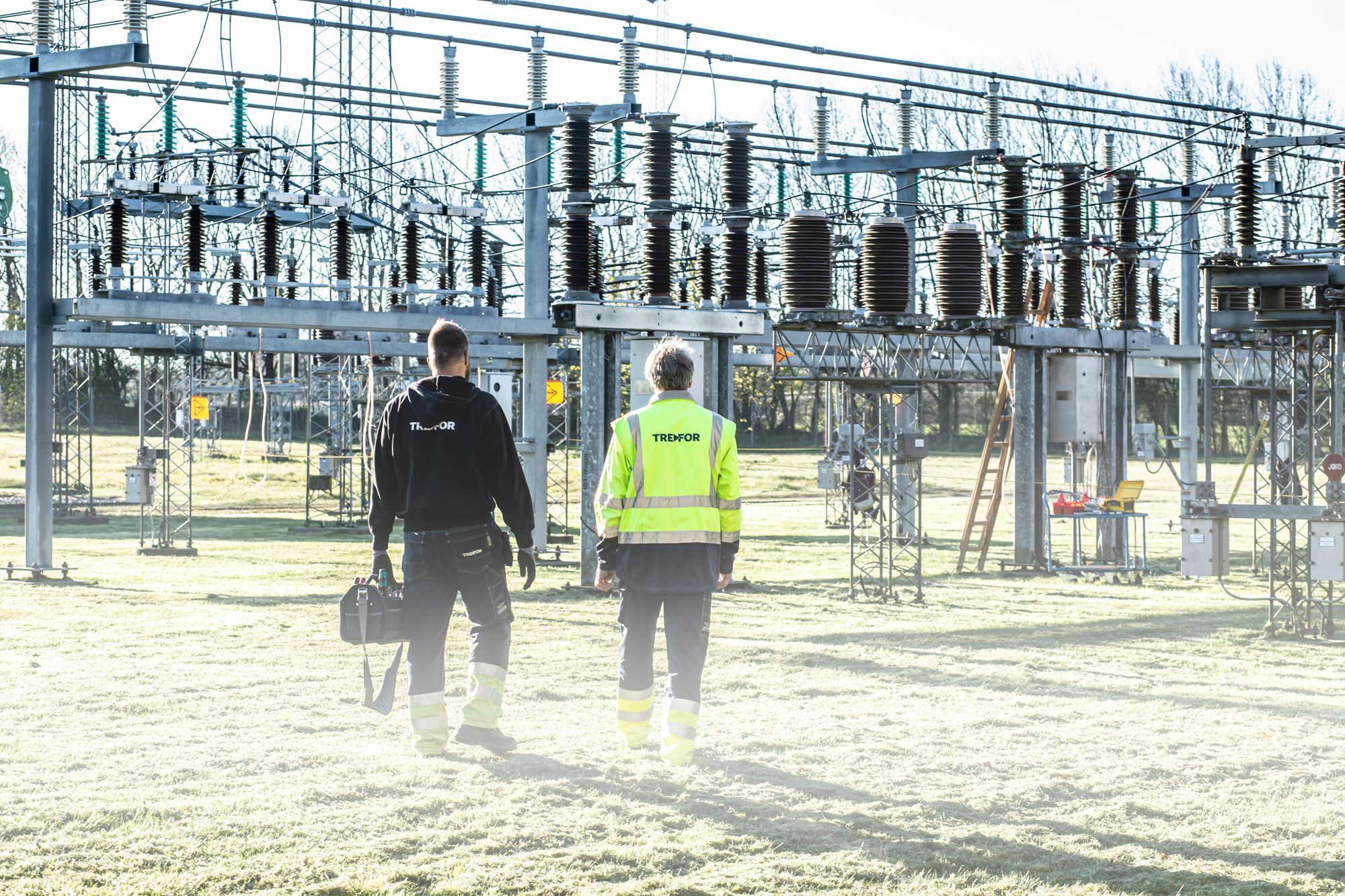 Two workers in safety gear walking toward high-voltage equipment at a substation